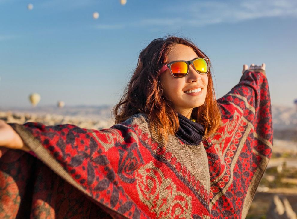 Donna sorridente con mantello rosso, braccia aperte, mongolfiere sullo sfondo al tramonto.
