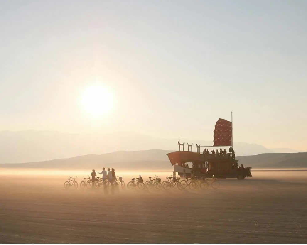 Tramonto nel deserto, biciclette e un carro con bandiera rossa solcano la polvere.